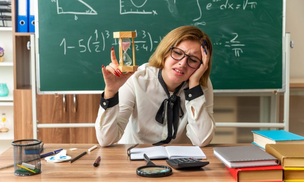 sad-with-closed-eyes-young-female-teacher-sits-table-with-school-supplies-holding-hourglass-classroom-min-scaled-1-1-1-1024x650-1.jpg