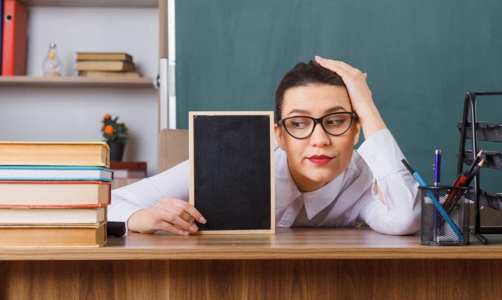 young-woman-teacher-wearing-glasses-with-small-chalkboard-sitting-school-desk-front-blackboard-classroom-looking-tired-overworked-min-scaled-2-2-1024x683-1.jpg